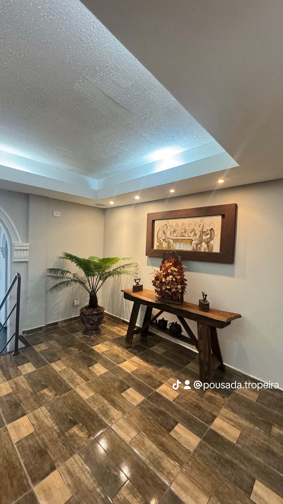 A well-lit entryway features a wooden table adorned with decorative elements and a potted plant. The floor is tiled in warm, earthy tones, and the ceiling is elegantly designed with recessed lighting, creating a welcoming atmosphere.