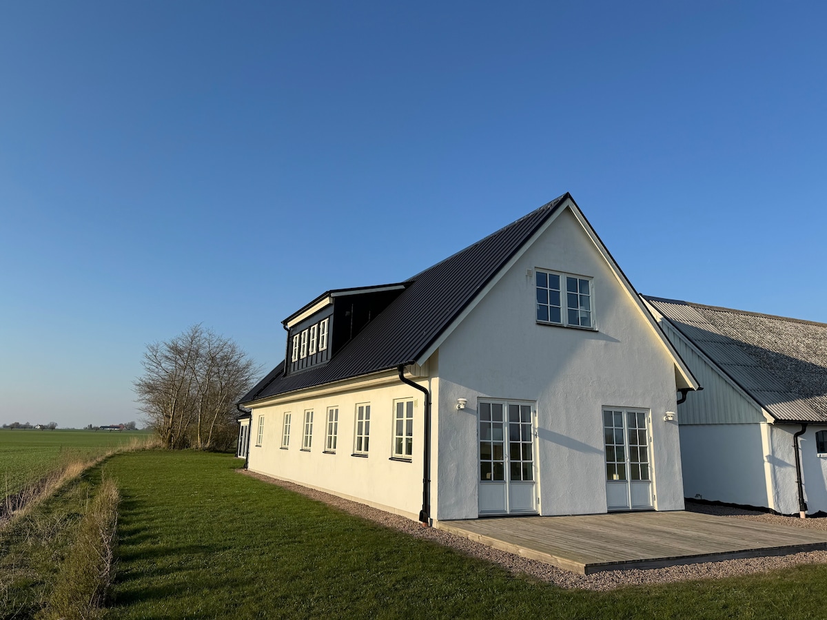 A white, two-story building features large windows that allow natural light to fill the interior. The structure is surrounded by green grass and has a wooden deck extending from the front. The clear blue sky complements the rural setting.