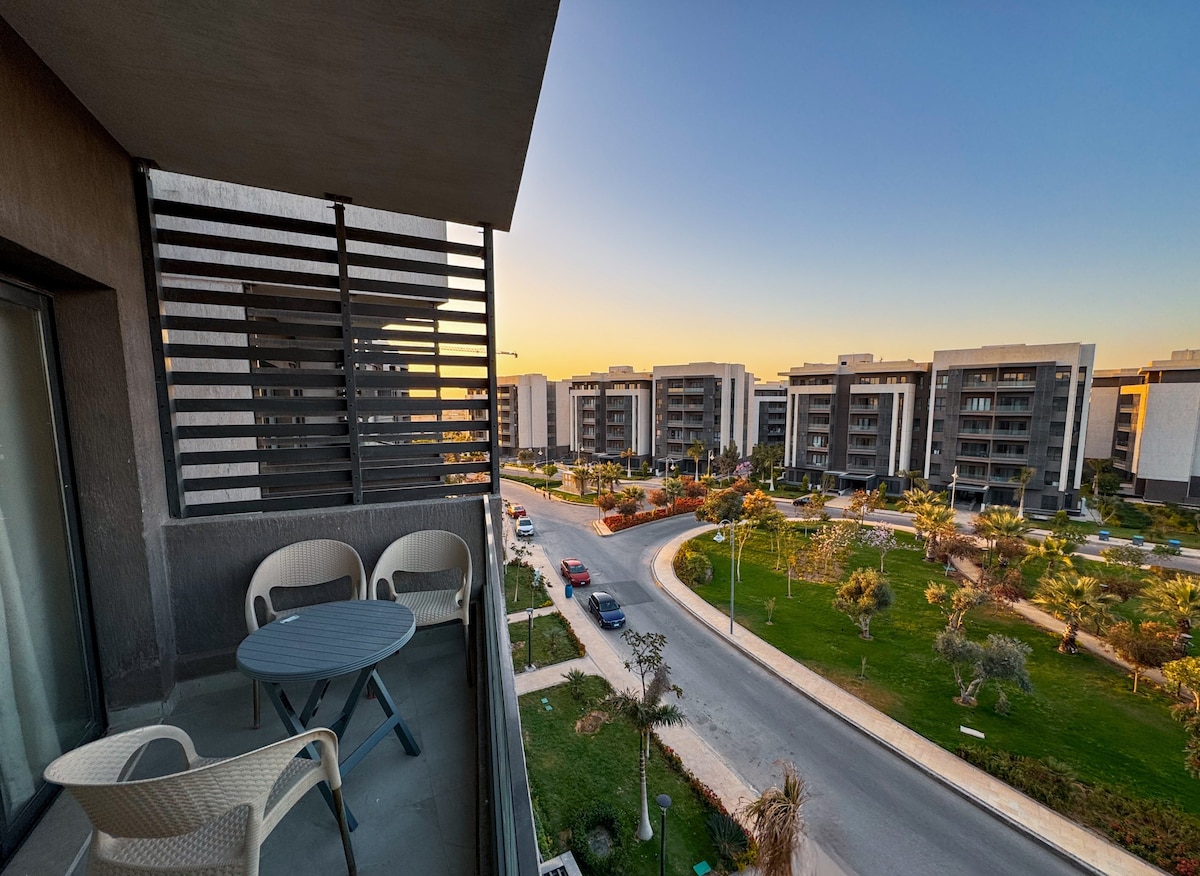 A balcony offers a view of the surrounding landscaped grounds, showcasing well-maintained lawns and modern buildings. A small table and two chairs are positioned on the balcony, providing a place to enjoy the sunset or fresh air.