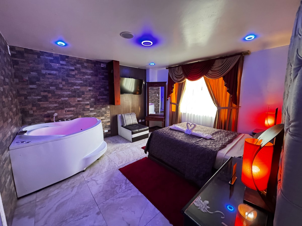 A spacious room features a large circular bathtub and a comfortable dark-colored bed with decorative pillows. Subtle lighting enhances the ambiance, while a wall-mounted television provides entertainment. A large window is covered by elegant drapery, letting in natural light.