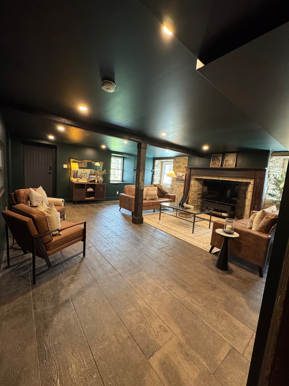 A cozy living room is depicted, featuring two armchairs and a loveseat arranged around a central coffee table. A stone fireplace serves as a focal point, while natural light filters through the windows, illuminating the wood flooring and dark ceiling.