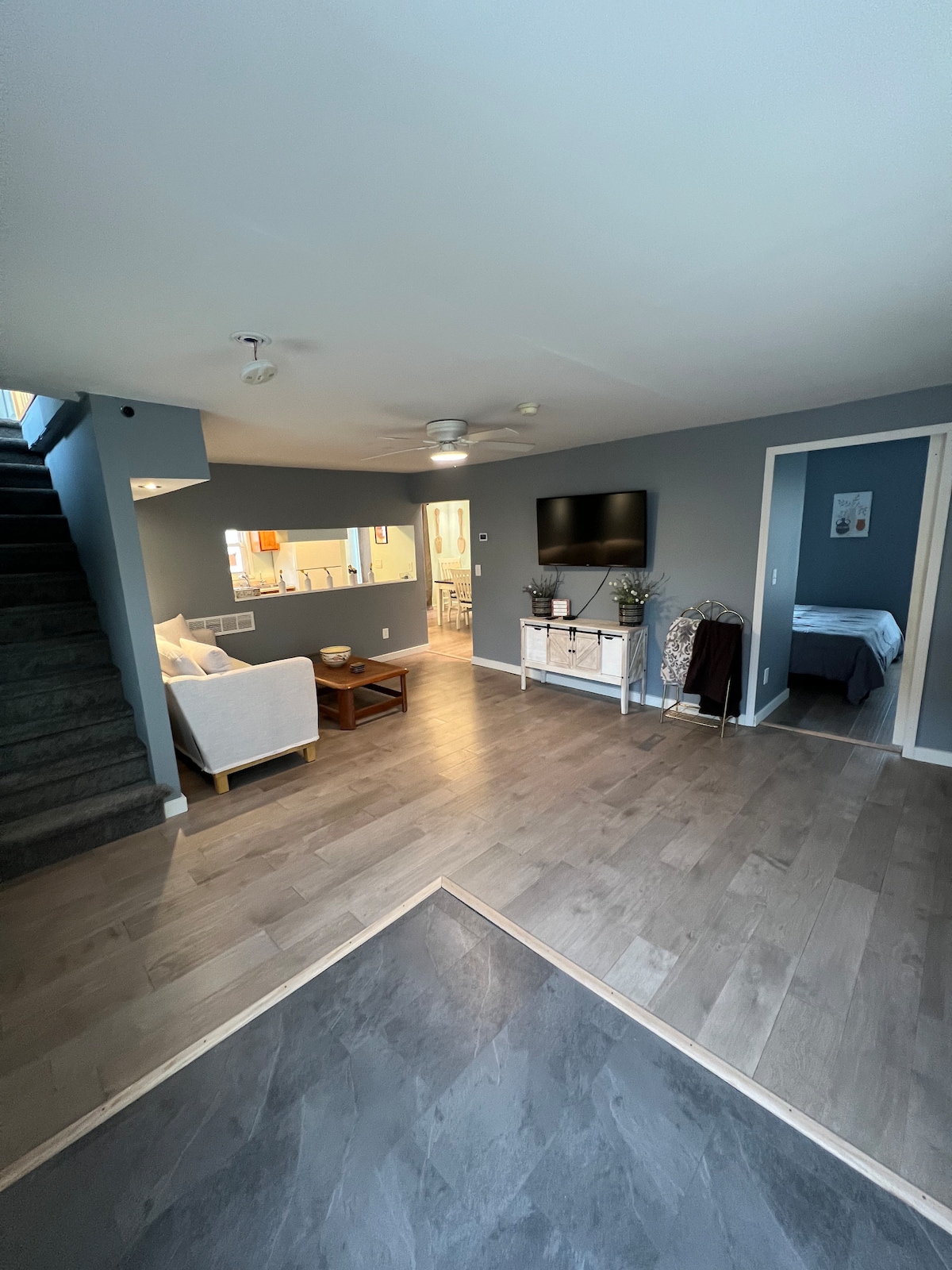 A spacious living area is visible, featuring a light-colored sofa and a wooden coffee table. A wall-mounted TV adds functionality, while a stairway leads to an upper level. The room is adorned with neutral wall colors, and a doorway in the background suggests access to additional bedrooms.