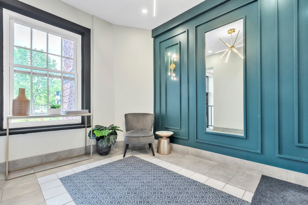 An inviting entrance area features a green accent wall, complemented by a large mirror and modern light fixture. A gray armchair is positioned beside a small side table. A decorative plant is placed in the corner, enhancing the overall ambiance of the space.