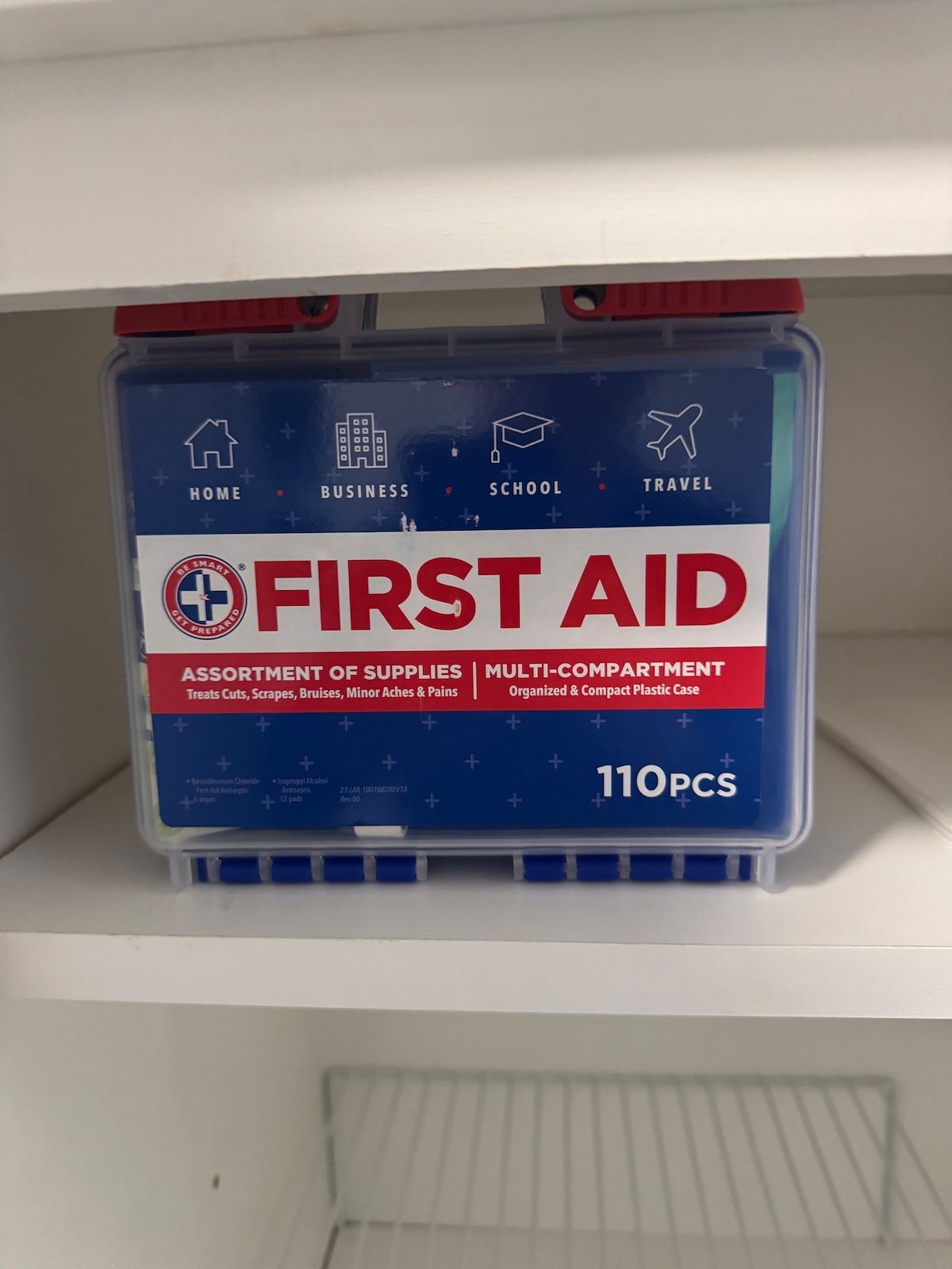 A clear plastic first aid kit is displayed on a shelf. The kit is labeled 'FIRST AID' and features icons representing home, business, school, and travel. Inside, a variety of medical supplies are organized within multiple compartments, totaling 110 pieces.