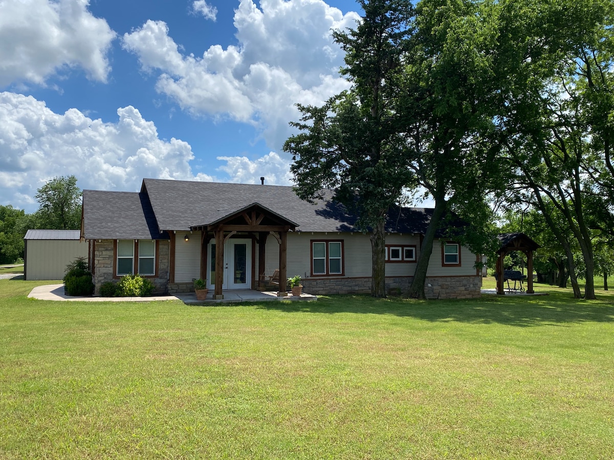 A single-story home is set within a green landscape, surrounded by trees. The structure features a dark roof, stone accents, and large windows. Sunlight brightens the exterior, reflecting a welcoming entryway and a spacious lawn leading to the front door.
