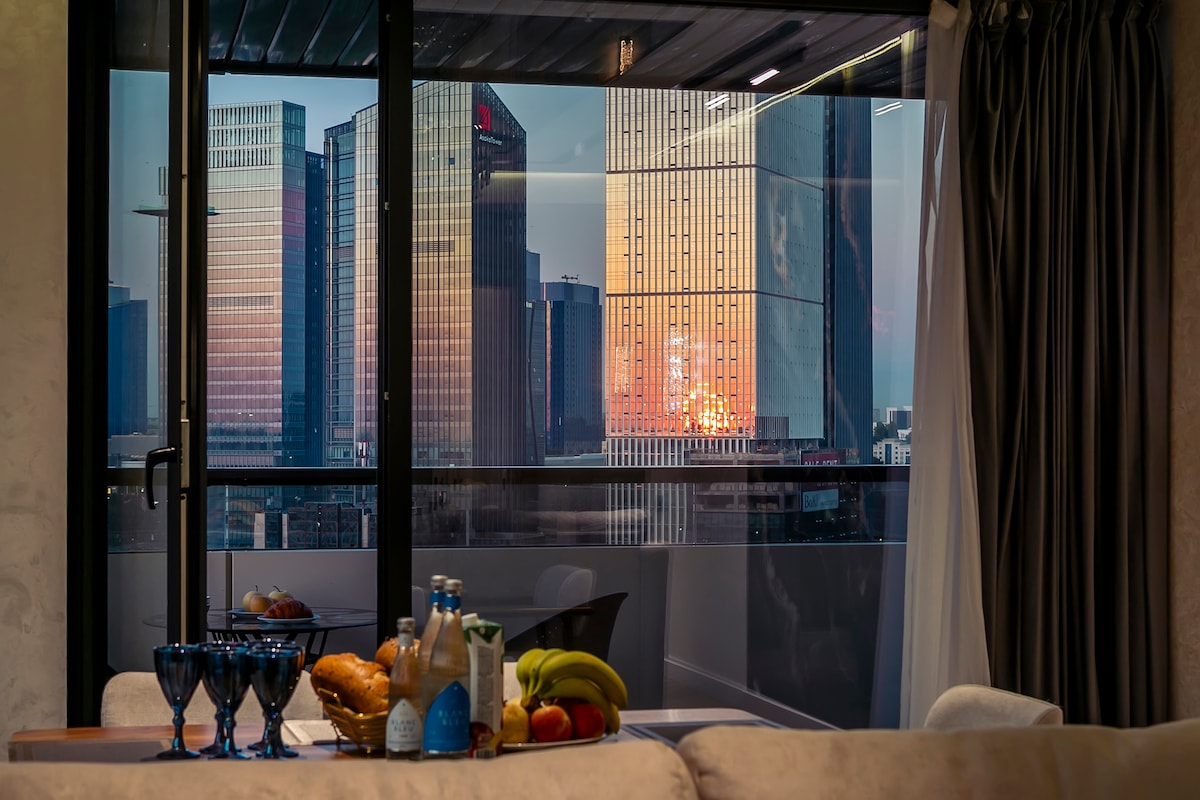 A dining area is visible, featuring a wooden table set with three glasses, a bottle, and a selection of fruits. The background displays a panoramic view of modern skyscrapers reflecting warm sunset hues through floor-to-ceiling glass doors.