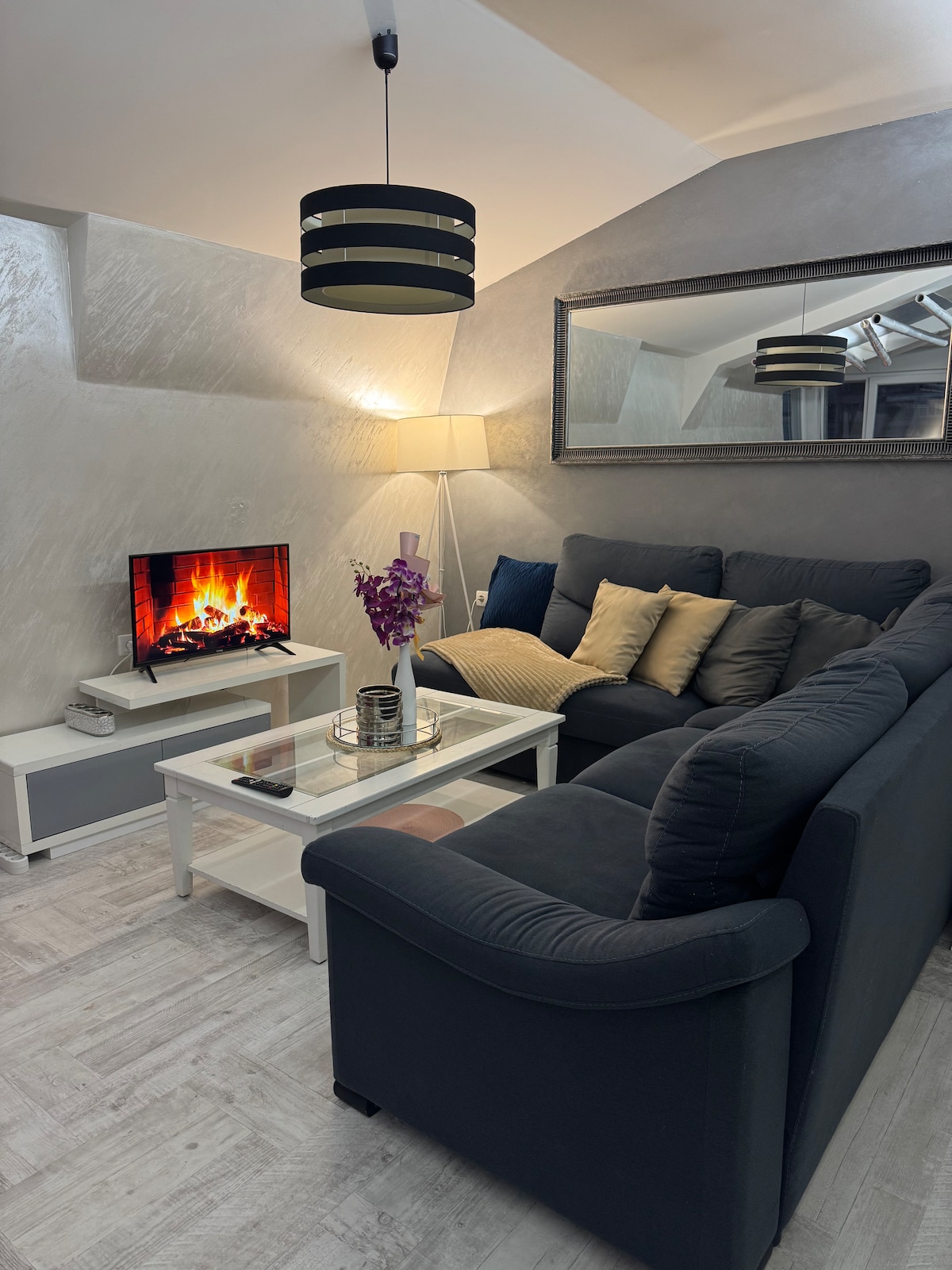A cozy living area is presented, featuring a dark sofa adorned with decorative pillows. A modern coffee table sits in front, topped with a vase of flowers. A television displays a virtual fireplace, and soft lighting is provided by a floor lamp and pendant light.