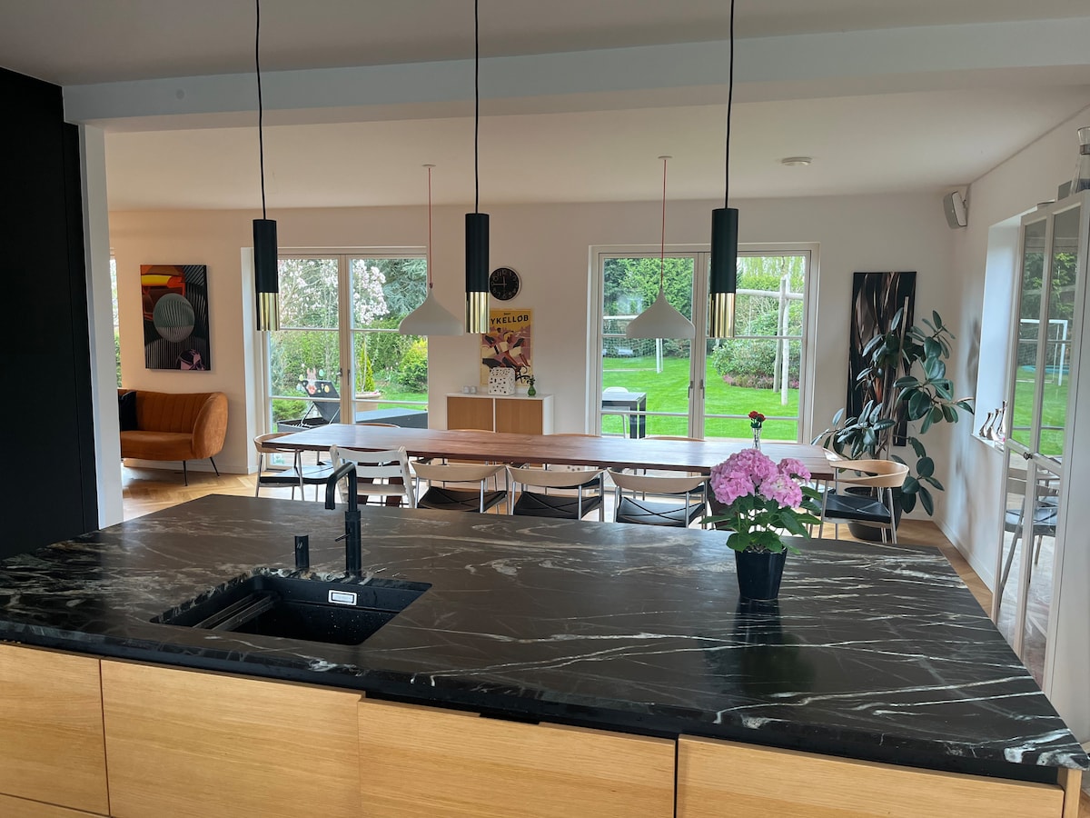 An open kitchen area features a dark marble countertop with a small potted plant. Large windows provide views of the garden, while a dining table is positioned in the background, surrounded by light-colored chairs. Soft lighting hangs above the kitchen island.