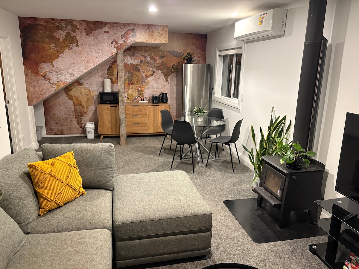 A cozy living area features a large sectional sofa with a yellow accent pillow. A wall adorned with a world map serves as a backdrop to a dining space with a round table and four chairs. A modern kitchenette is visible, alongside a sleek grey fridge.