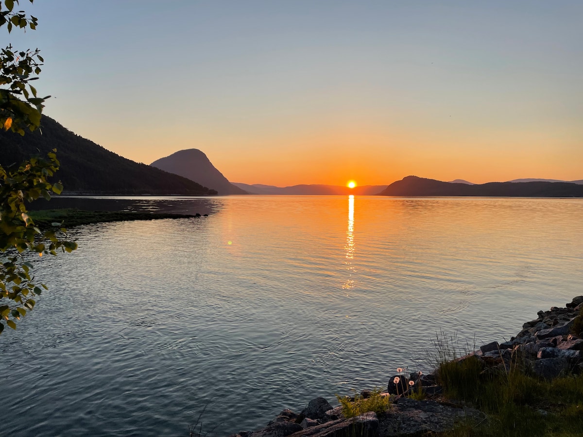 The image captures a serene waterfront view during sunset, with the sun's rays reflecting on calm waters. Mountains frame the scene in the background, while the shoreline features a rocky edge and some green vegetation, creating a tranquil atmosphere.