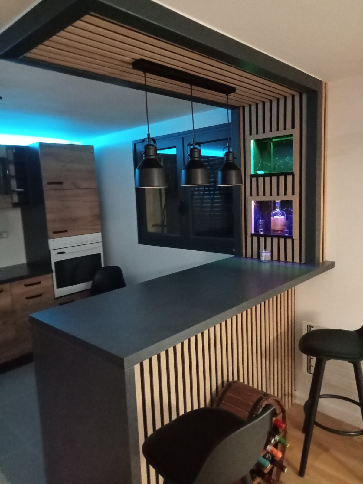 A modern kitchen area features a minimalist bar with three pendant lights hanging overhead. Behind the bar, wooden slats frame a decorative niche with green and purple accent lighting. Kitchen cabinetry in natural wood tones complements the sleek countertop.