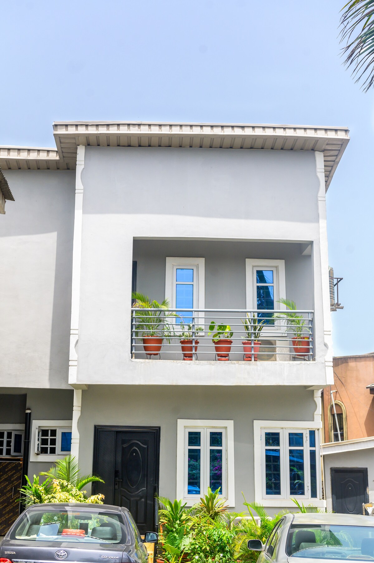 A modern two-story building is featured, highlighted by a balcony adorned with potted plants. Large windows are framed in blue, allowing natural light to brighten the facade. The entryway is accentuated by a dark door, surrounded by a lush garden featuring greenery.