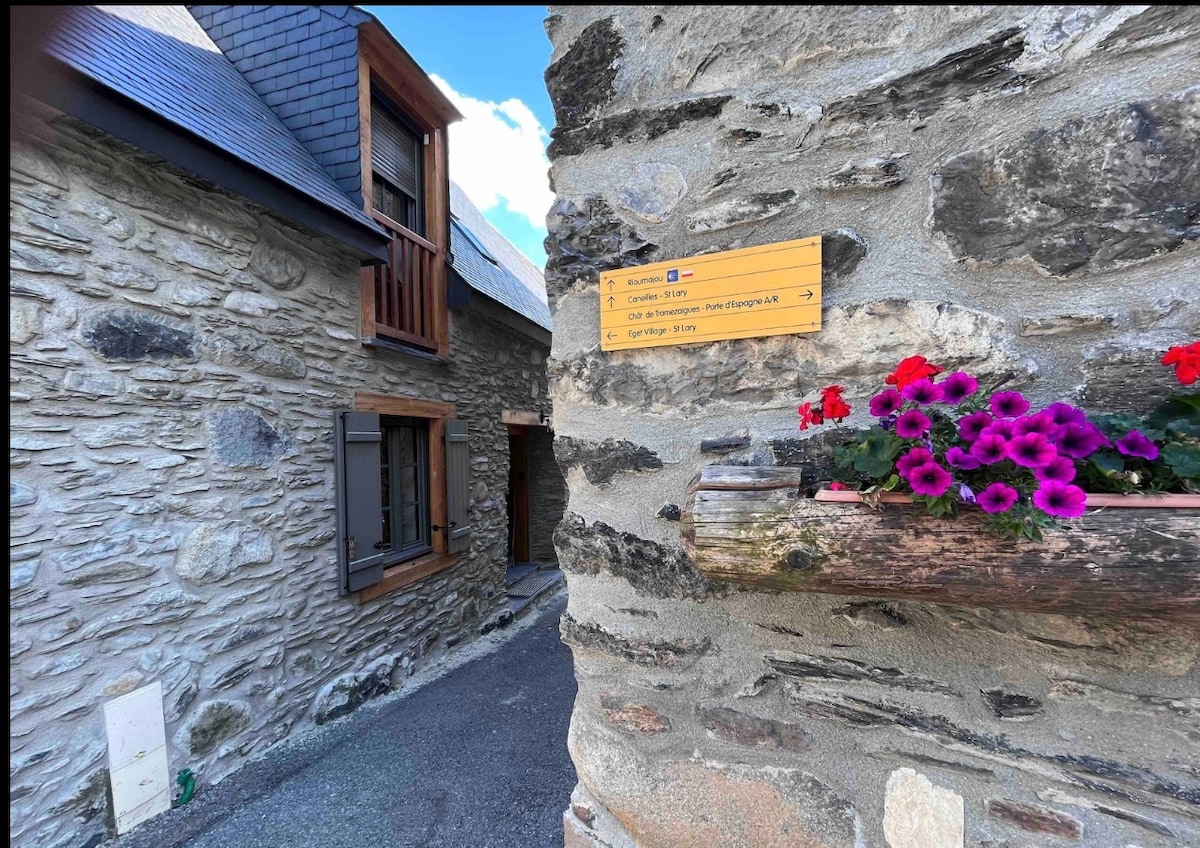 A rustic stone façade of the property is displayed, complemented by a wooden sign indicating directions. Bright purple flowers are seen in a planter, adding a touch of color to the entrance while the surrounding natural stone conveys the building's traditional charm.