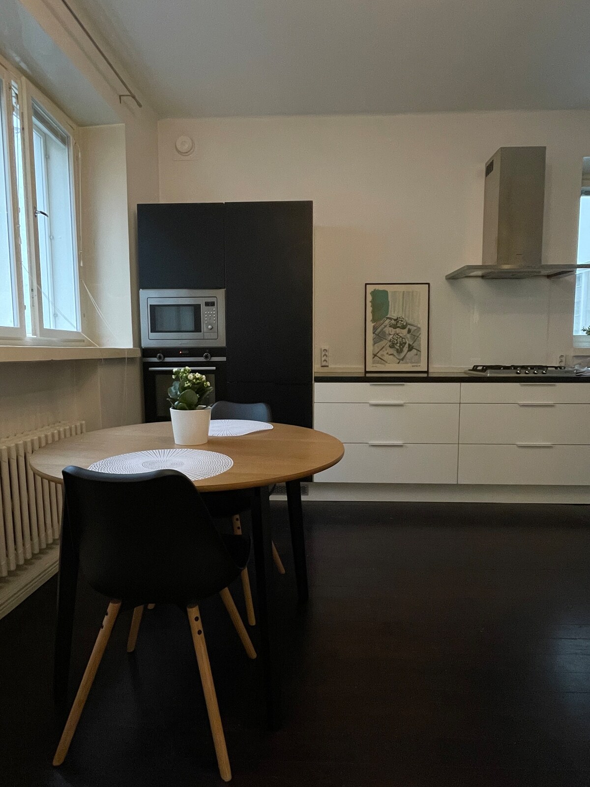 A well-equipped kitchen features modern appliances, including a microwave and stovetop. A round dining table is surrounded by four black chairs. Natural light enters through large windows, illuminating the space. Minimalist décor is complemented by a potted plant on the tabletop.
