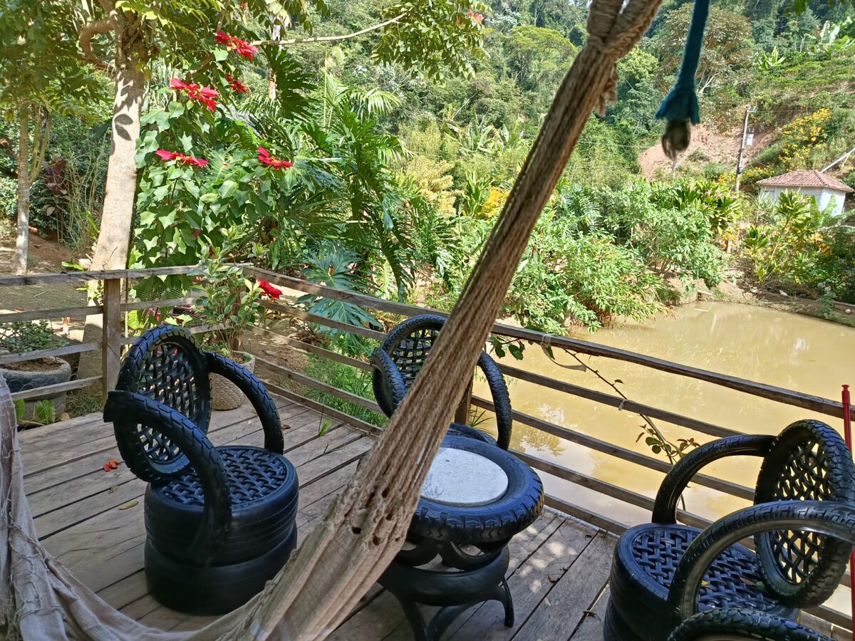 An outdoor seating area features several round tables surrounded by black wicker chairs, overlooking a tranquil pond. Lush greenery and vibrant flowers provide a natural backdrop, creating a peaceful environment for relaxation. A wooden deck adds warmth to the setting.