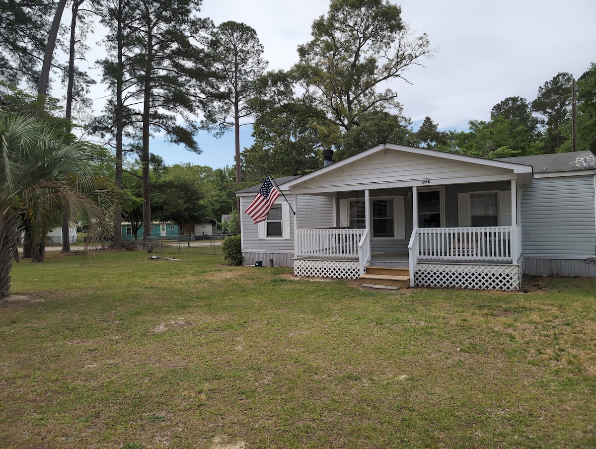 A charming single-story house is presented with a welcoming front porch. An American flag is displayed on the porch, surrounded by a spacious lawn. Tall pine trees and greenery enhance the outdoor setting, providing a peaceful atmosphere.