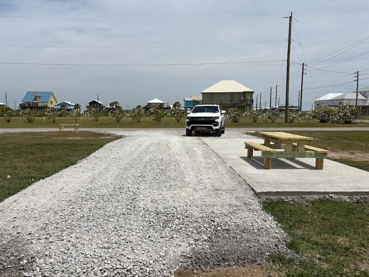 A gravel lot is featured, providing ample parking space for RVs or campers. A brand new wooden picnic table is positioned on a concrete slab, inviting gatherings. In the background, a mix of residential structures and lush greenery can be observed, enhancing the environment.