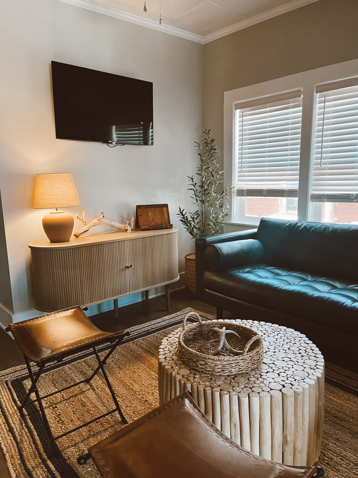 A comfortable living room is presented, featuring a blue leather couch and two folding chairs. A round coffee table made of stacked wood log segments is centered. A lamp and decorative elements, including a small basket, add subtle warmth. Natural light filters through the window blinds.