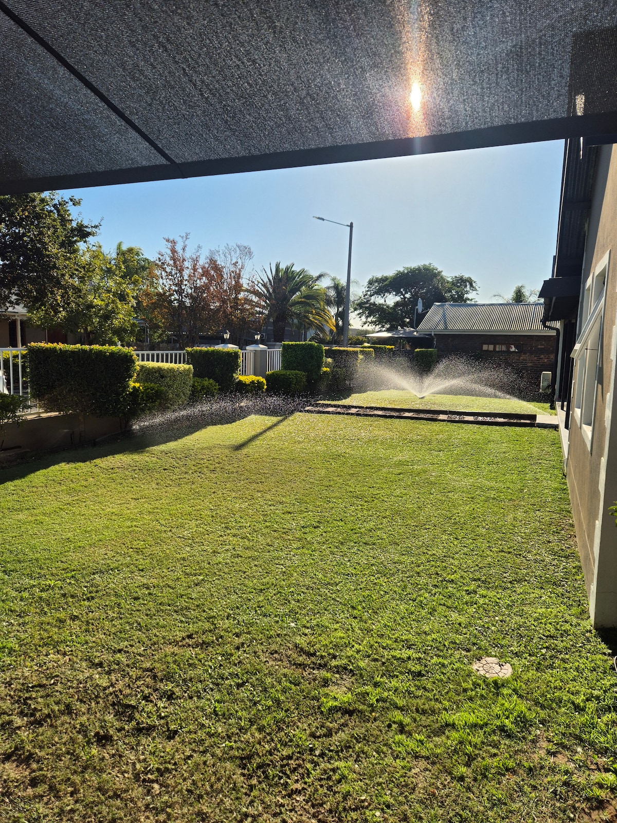 A well-maintained garden area features a lush green lawn, with sprinkler systems gently irrigating the grass. Surrounding shrubs and small trees add to the natural setting, while sunlight filters through a shaded canopy above. The scene captures a calm outdoor space ideal for relaxation.