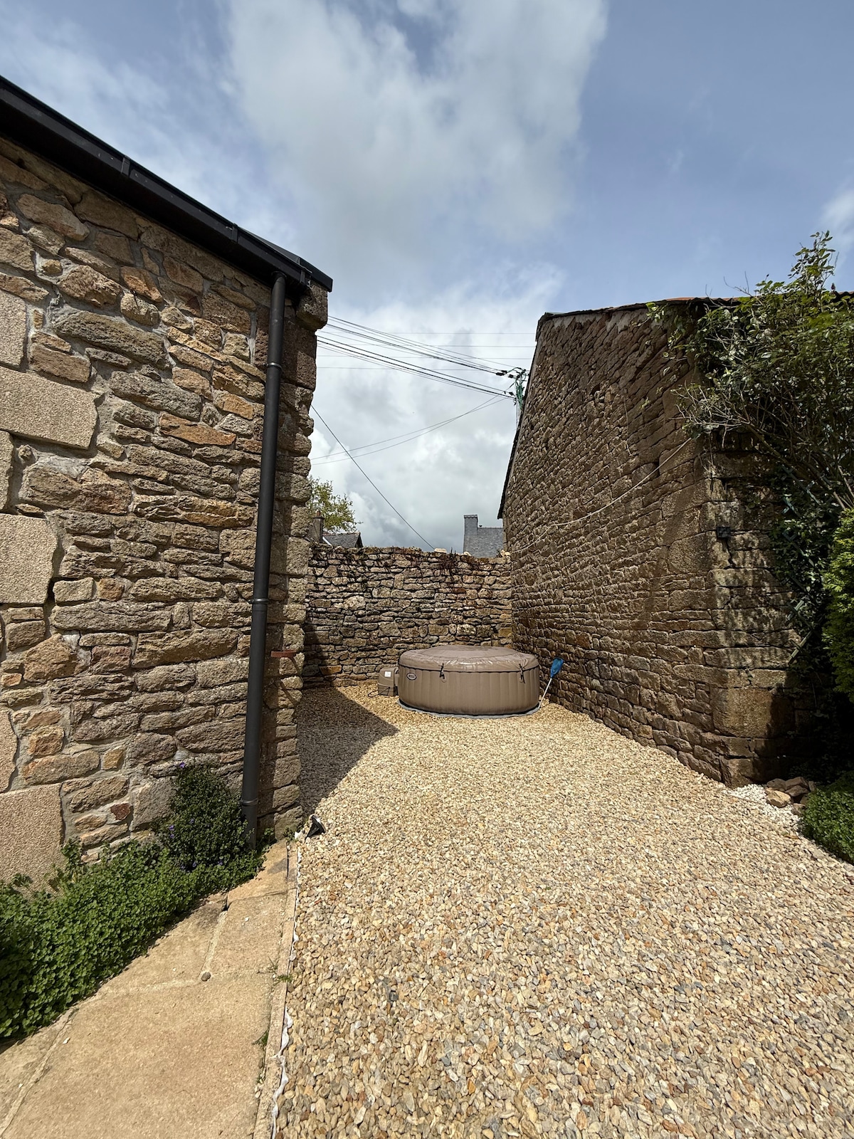 A gravel pathway leads to a circular hot tub, framed by stone walls on either side. Green foliage is present near the edges, providing a touch of nature. The sky is partly cloudy, contributing to the tranquil setting.