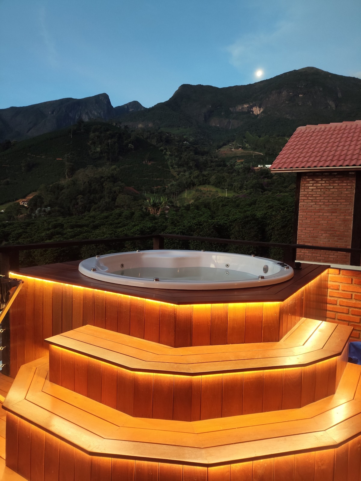 A hot tub is positioned on a wooden platform with illuminated steps, surrounded by lush greenery and mountainous terrain. The evening sky is visible above, featuring soft hues and a hint of moonlight.