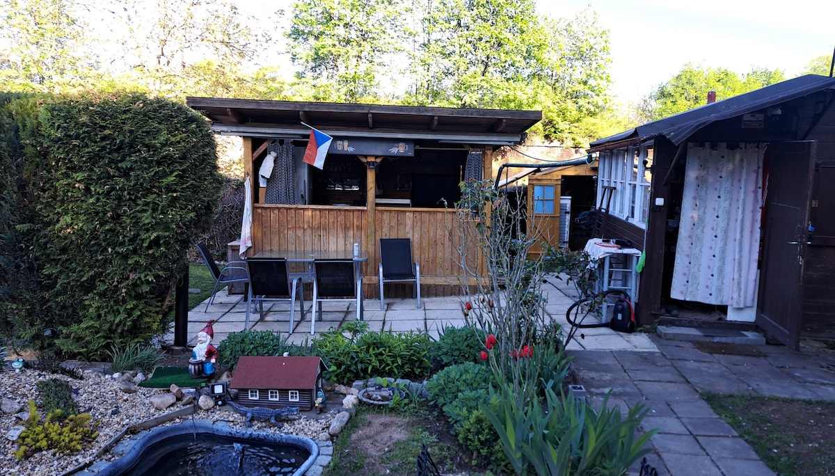 An outdoor area features a wooden gazebo with seating for four. A garden with various plants, including flowers and greenery, surrounds a decorative pond. A small house-shaped decoration is situated in the garden, and a storage shed is located to the right.