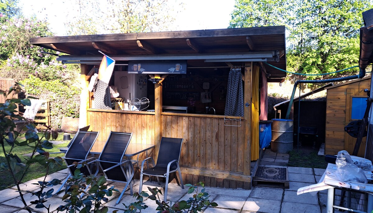 A charming outdoor structure features wooden paneling and a sloped roof. Two lounge chairs are positioned on a stone patio, while colorful fabrics drape around the bar area. Lush greenery surrounds the space, creating a serene environment, ideal for relaxation.