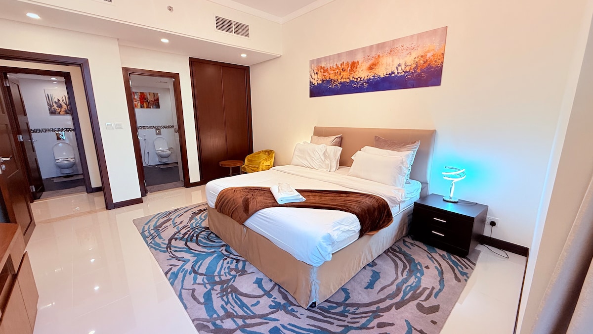 A spacious bedroom features a large bed with white linens and a brown throw. An abstract artwork is displayed above the bed. A small bedside table holds a modern lamp, and a plush chair in the corner adds comfort. Two doors lead to bathroom facilities in the background.