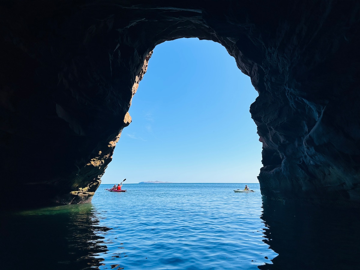 A natural rock formation creates a striking archway, framing a serene view of the vibrant blue ocean. Two kayakers are seen paddling leisurely, their colorful kayaks contrasting against the calm water and dramatic cliffs.