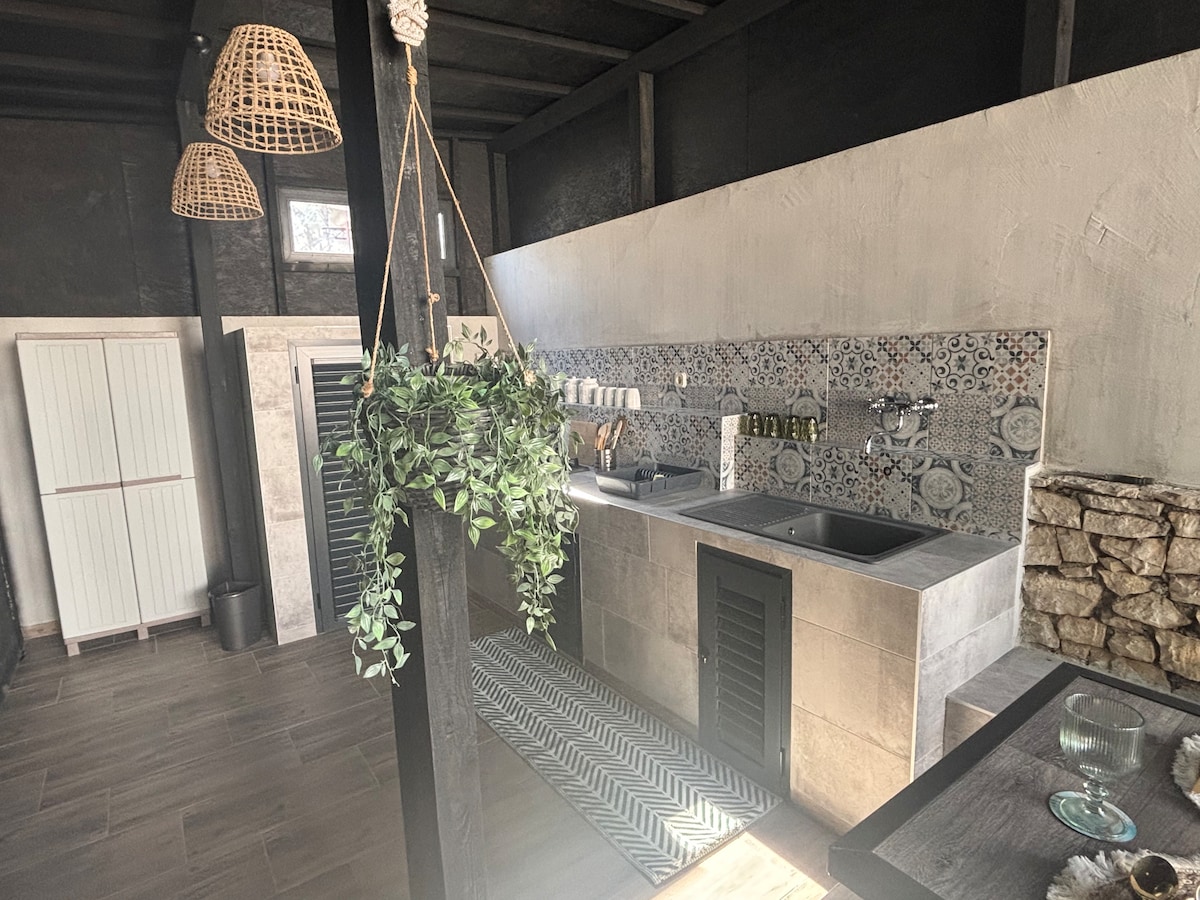 An open-plan kitchen features modern appliances and a decorative tile backsplash. A hanging plant adds a touch of greenery, while woven pendant lights provide soft illumination. Stone walls and light wood accents create a warm environment, inviting culinary creativity.