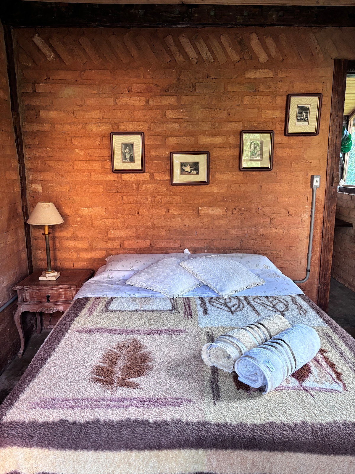 A cozy sleeping area features a spacious bed adorned with a patterned blanket and neatly arranged towels. Soft lighting from a bedside lamp illuminates exposed brick walls and framed photographs, creating a soothing and comfortable ambiance.