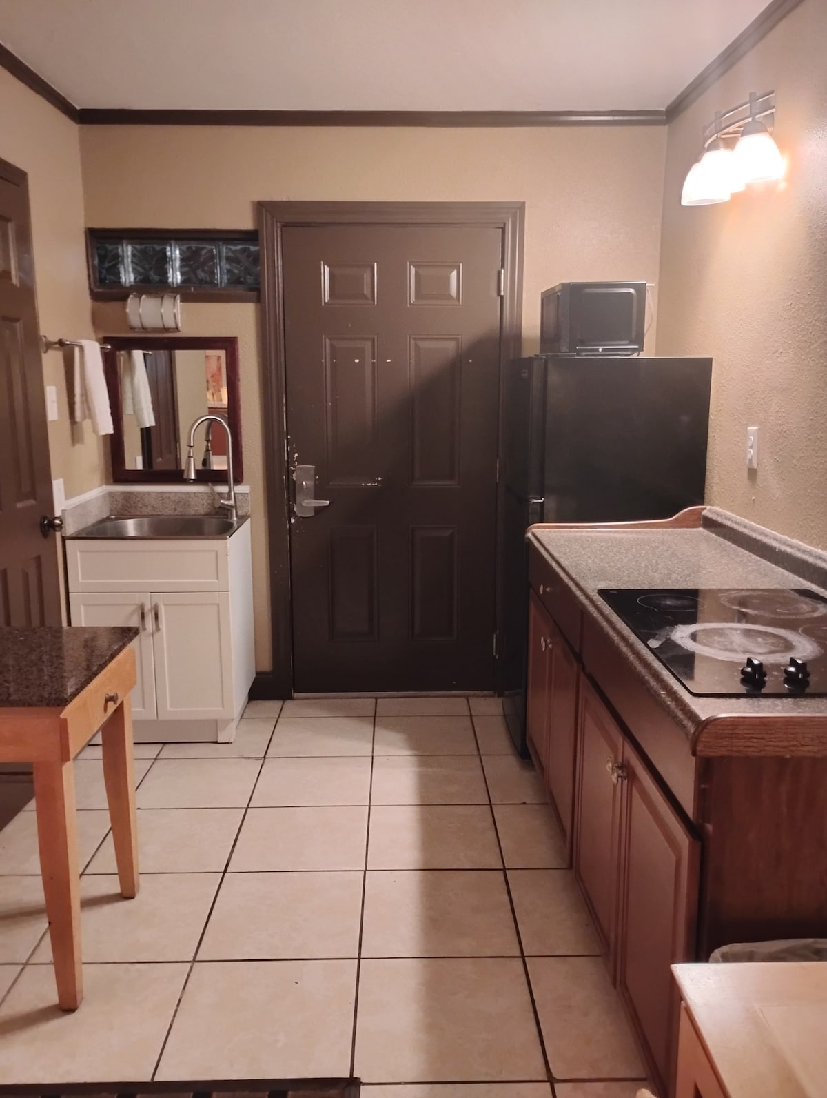 The kitchen area includes a dark wood countertop and cabinets. A compact refrigerator and microwave are positioned next to the entry door. A small wooden table is situated near the entrance. The tiled floor adds a practical touch to the space.