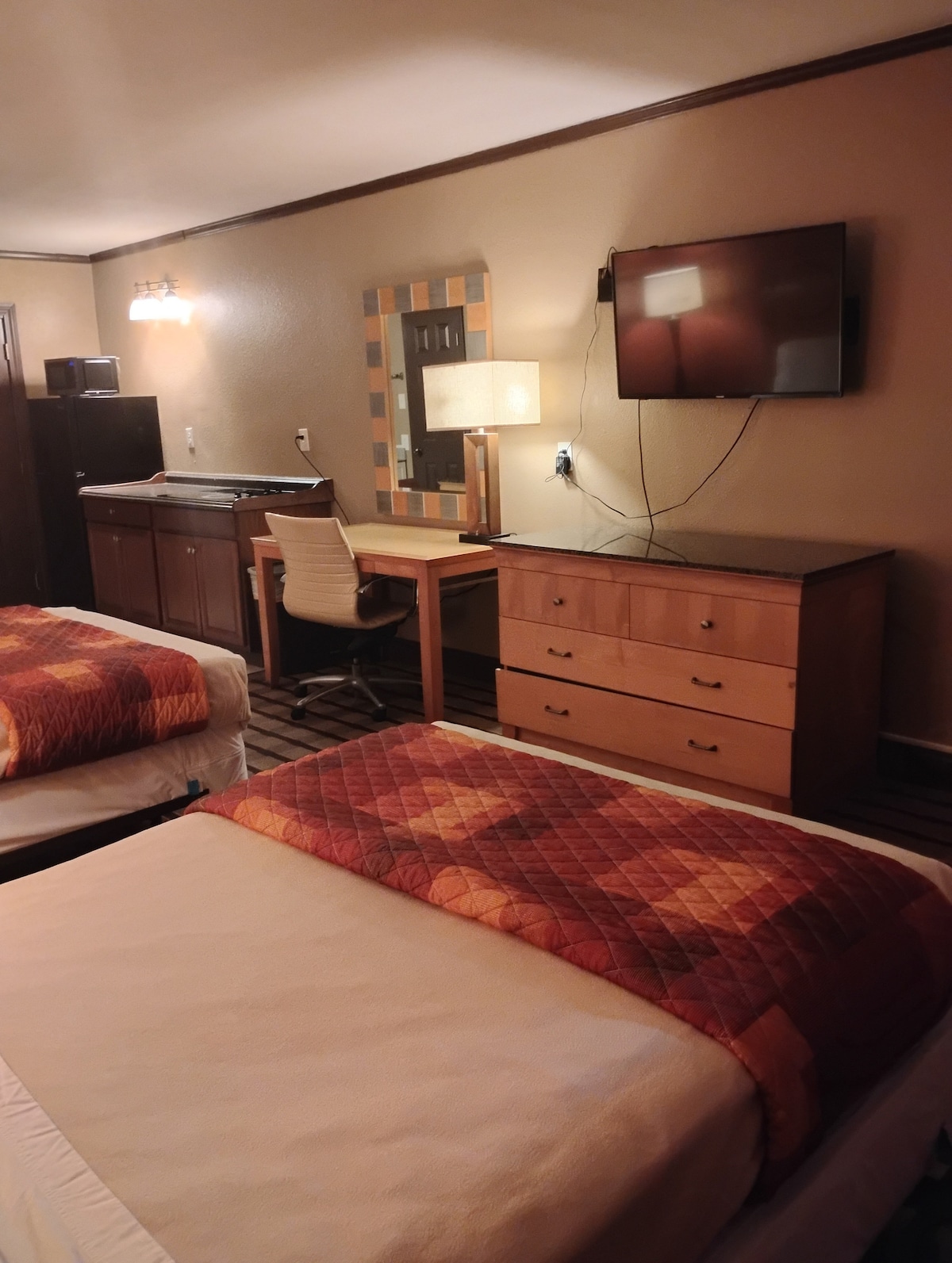 The room features two beds with light-colored linens and patterned throw blankets. A wooden dresser and desk provide storage and workspace. A flat-screen TV is mounted on the wall. An additional kitchenette area is equipped with cabinetry, enhancing functionality.