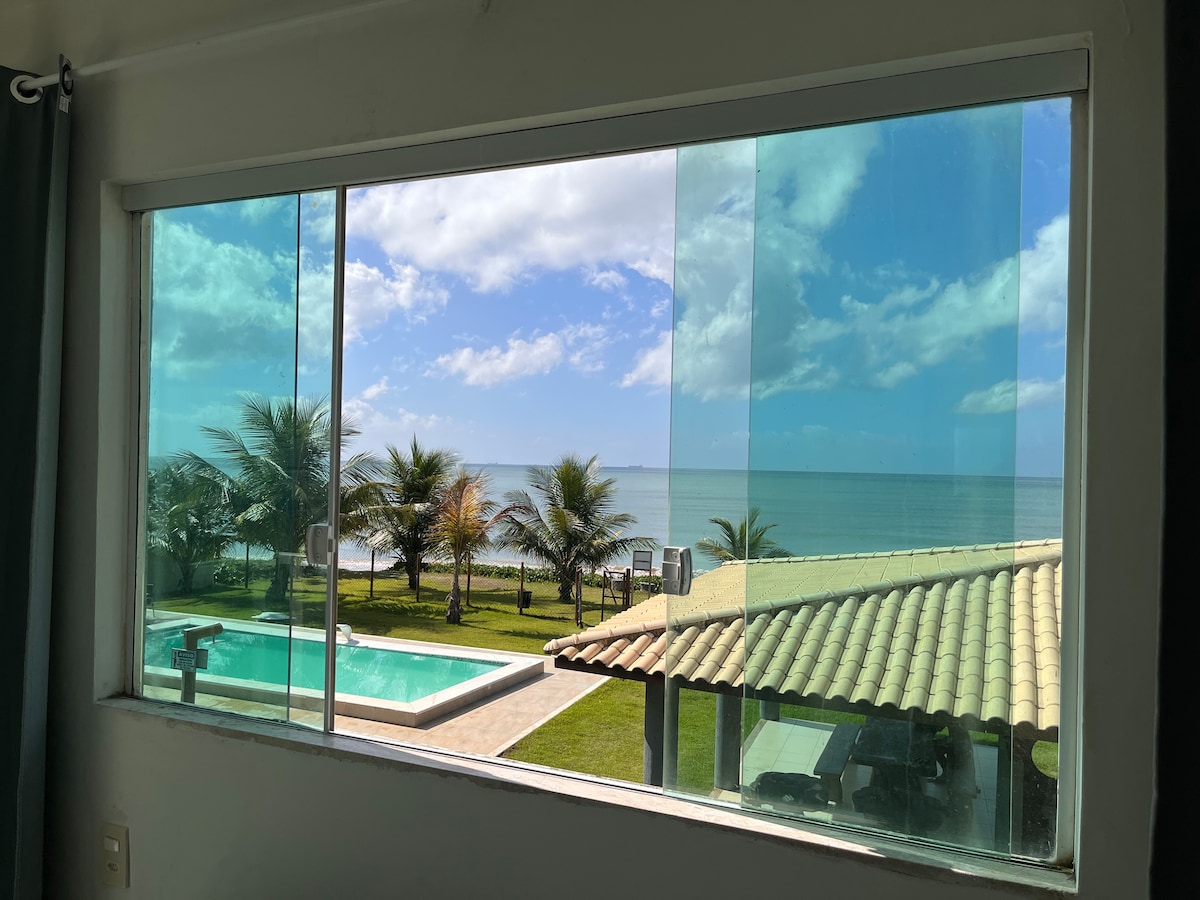 A panoramic view is captured through a large window, showcasing the serene ocean and lush palm trees. The inviting blue pool glistens in the sunlight, complementing the vibrant greenery of the garden below.