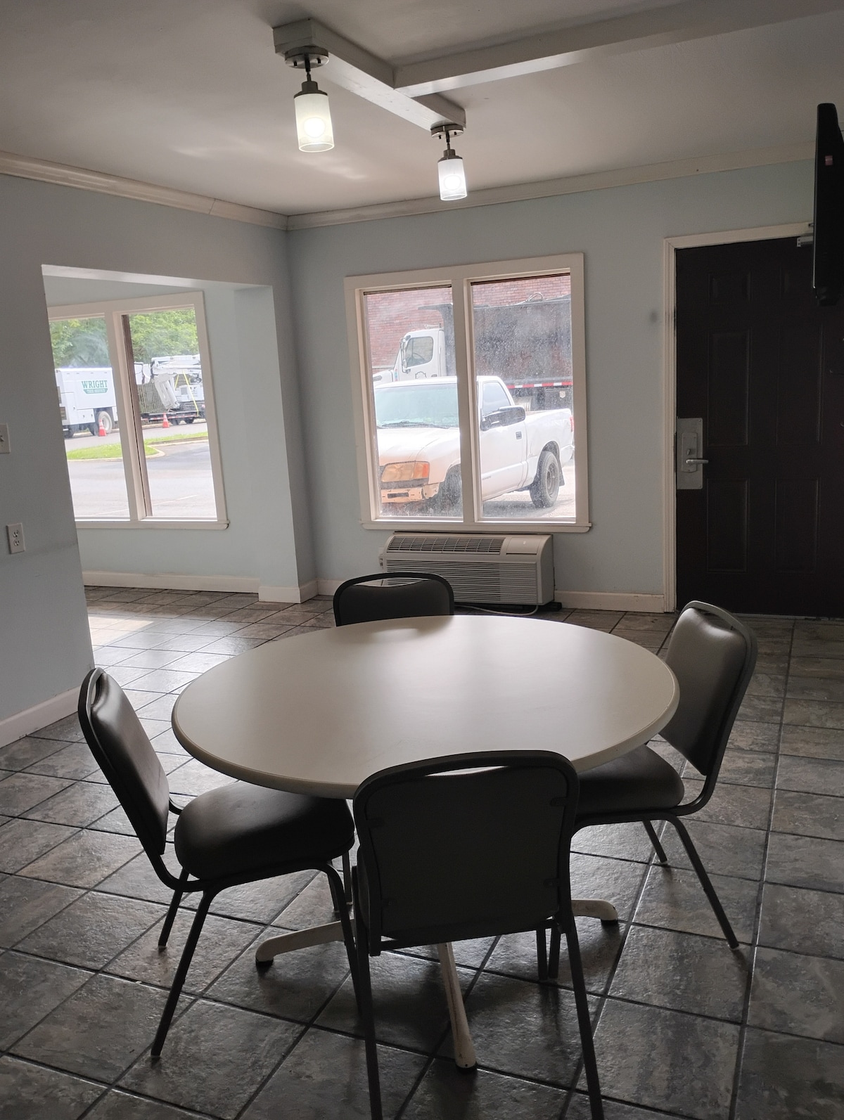 A dining area is shown, featuring a round table surrounded by four chairs. Light enters through two windows, offering views outside. The floor is tiled with a checkerboard pattern, and a dark door is visible on one side, leading to the apartment entrance.