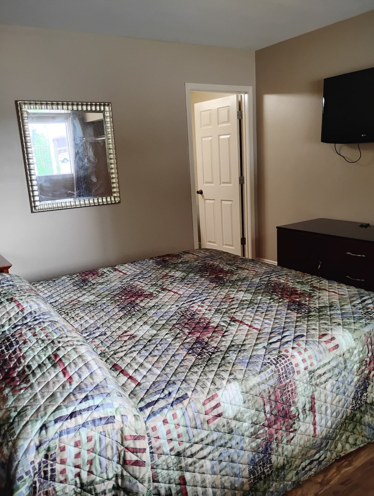 A spacious bedroom features a patterned quilt covering the bed, complemented by a mirror above a wooden dresser. A flat-screen TV is mounted on the wall. Light enters through a window next to the closed door, casting a soft glow in the room.