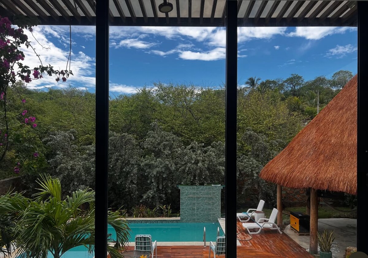 A view from inside showcases a swimming pool surrounded by lush greenery. A thatched-roof gazebo is visible nearby, along with lounge chairs positioned on a wooden deck. The sky is partly cloudy, adding depth to the serene outdoor setting.