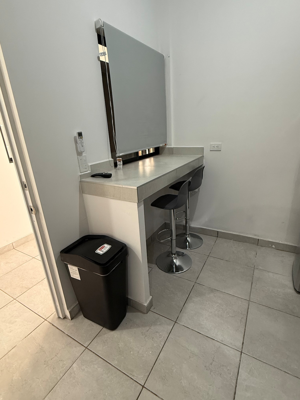 A small kitchenette area features a sleek bar counter with two modern stools. A wall-mounted mirror reflects the space, while a compact trash can is positioned beside the counter. The floor is covered with light-colored tiles, and the walls are painted in a soft shade.