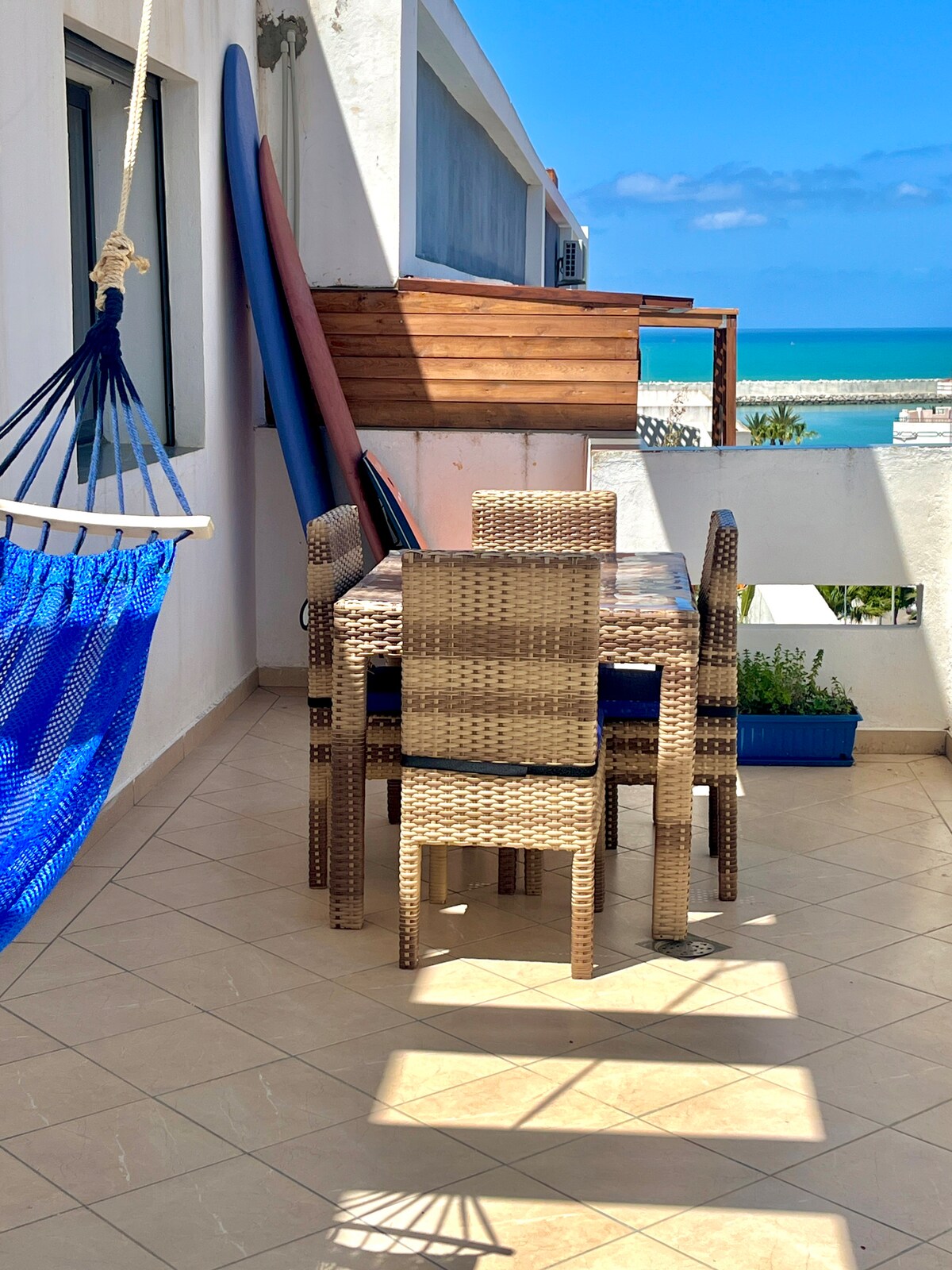 A terrace features a woven dining set with four chairs and a rectangular table, surrounded by natural light. A hammock is suspended nearby, and vibrant surfboards are propped against a wooden structure. The sea is visible in the background, enhancing the outdoor space.