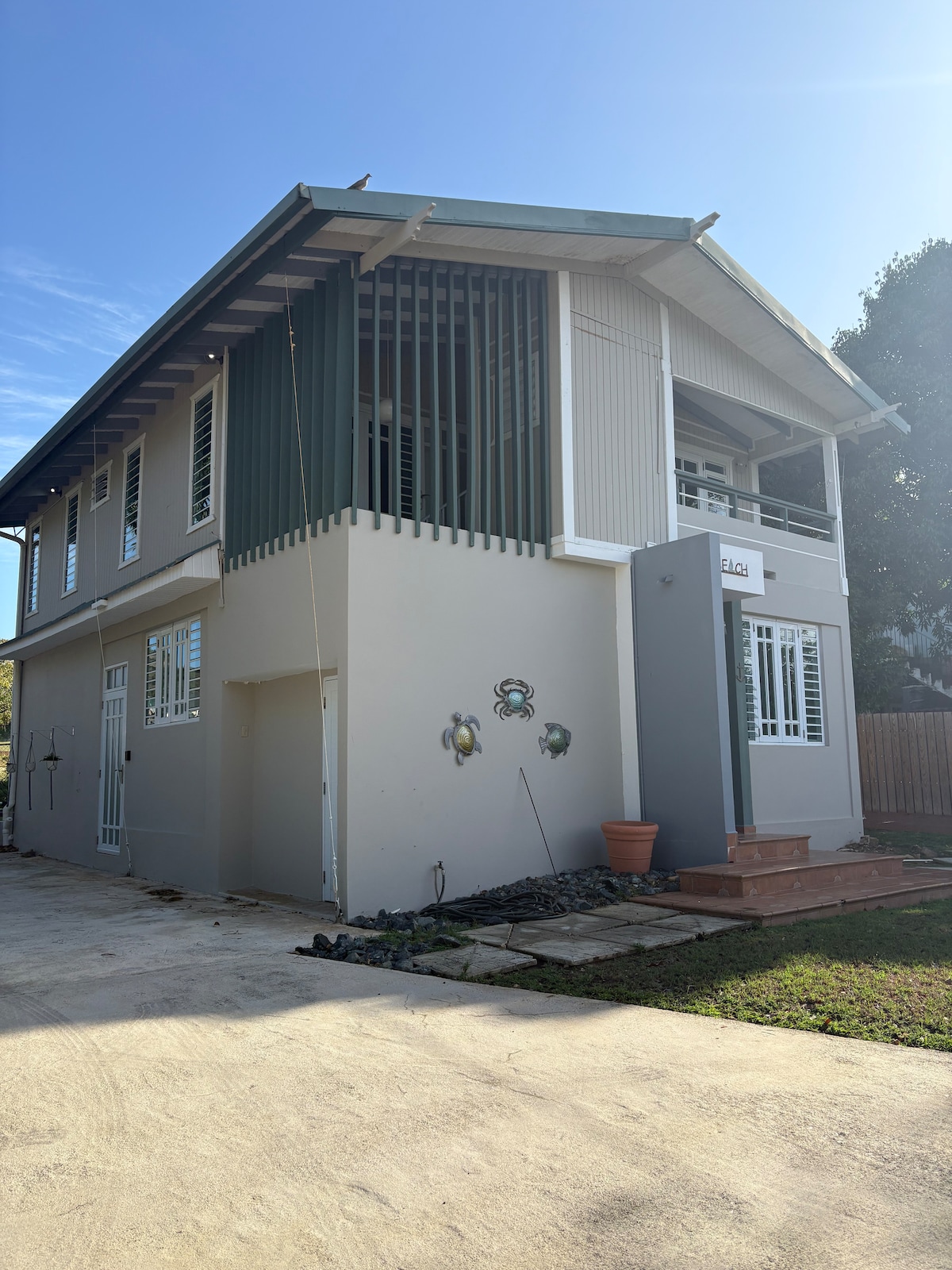A two-story building features a modern design with vertical wood slats on the upper level and a light grey exterior. The entrance is framed by a planter, and a concrete pathway leads to the steps, surrounded by green grass.