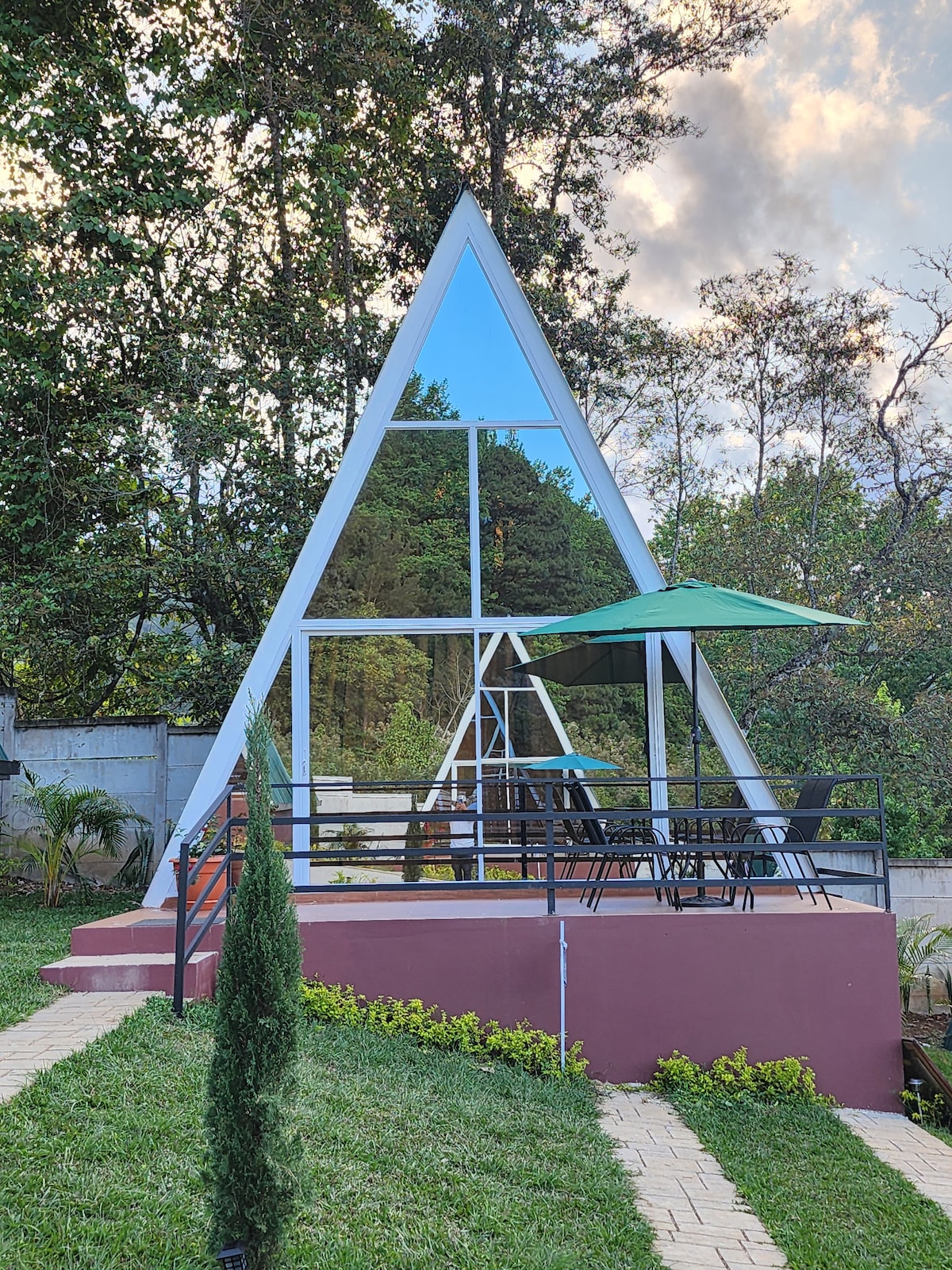A triangular-shaped cabin features large glass panels reflecting the surrounding greenery. A shaded outdoor area with a green umbrella is set up with table and chairs, providing an inviting space for relaxation. Pathways lined with greenery lead to the entrance.