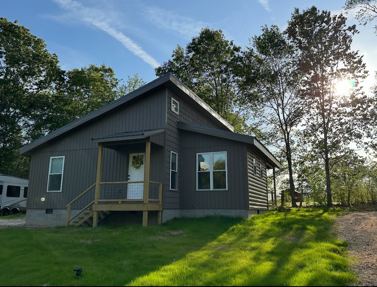 A rustic cabin is depicted, featuring a wooden front porch and symmetrical windows. The exterior is clad in dark brown wood, surrounded by green grass and trees. Sunlight filters through the leaves, illuminating the scene, suggesting a serene setting in nature.