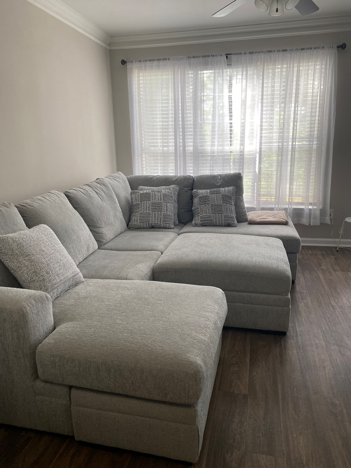 A light gray sectional sofa occupies a cozy corner of the room, adorned with two patterned throw pillows. Natural light filters through a large window, covered with sheer white curtains, illuminating the soft upholstery and wooden floor beneath.