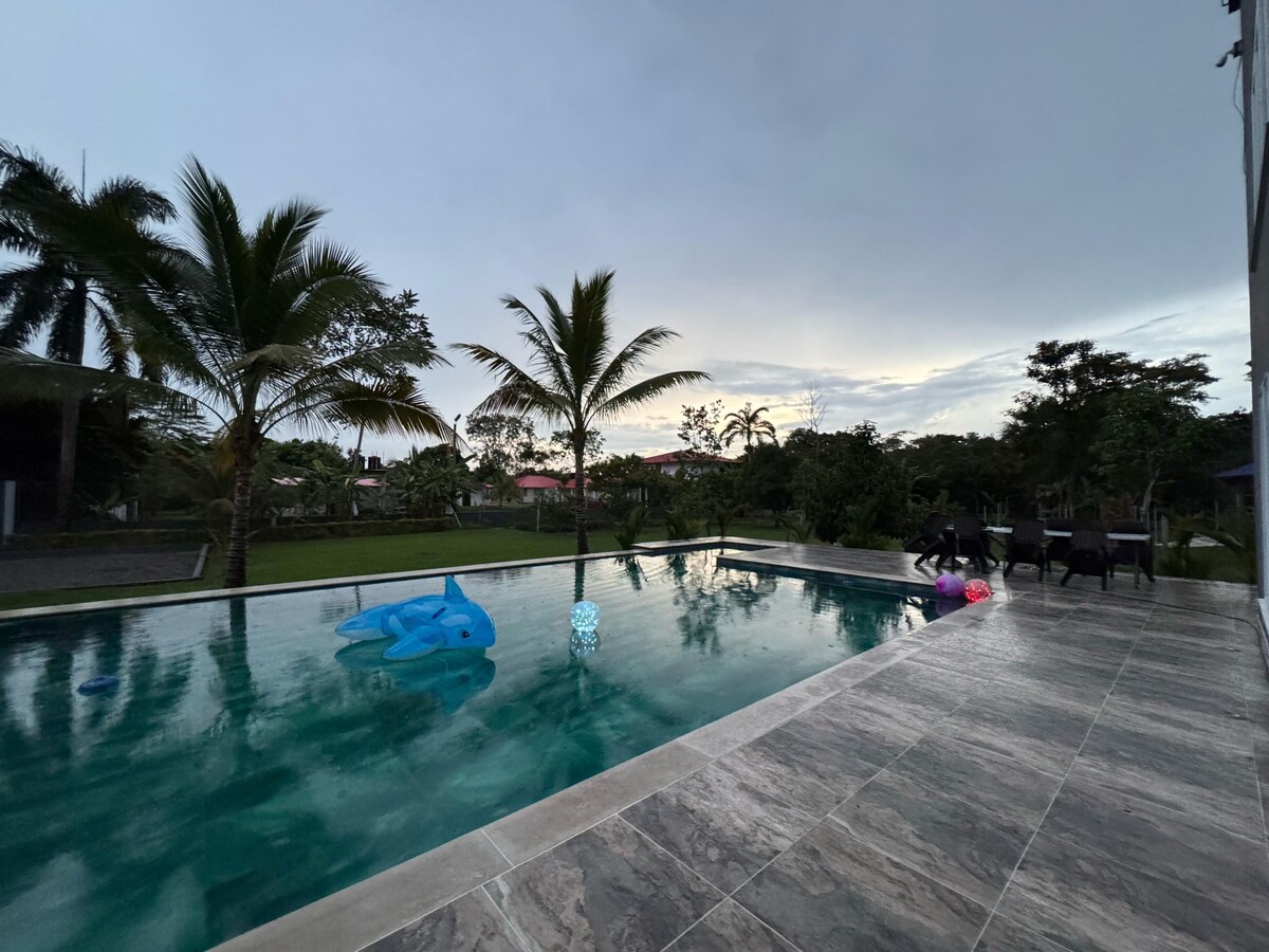 A serene pool area is bordered by lush greenery and palm trees. Colorful inflatable toys float peacefully on the water, while a paved deck lined with chairs offers a cozy space to relax. The overall setting provides a calm atmosphere, perfect for group gatherings.