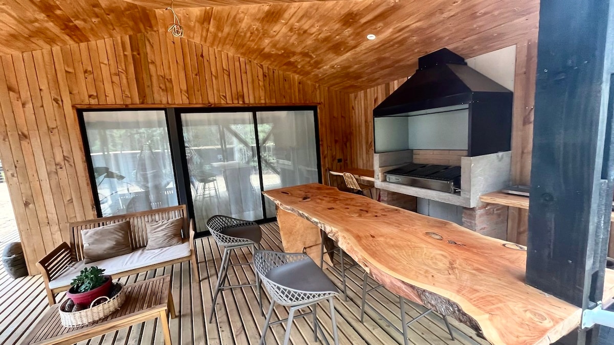 An outdoor dining area features a long wooden table crafted from natural wood. Modern chairs surround it, and a built-in barbecue is visible in the background. Large glass doors provide access to the interior space, enhancing the connection with the natural surroundings.