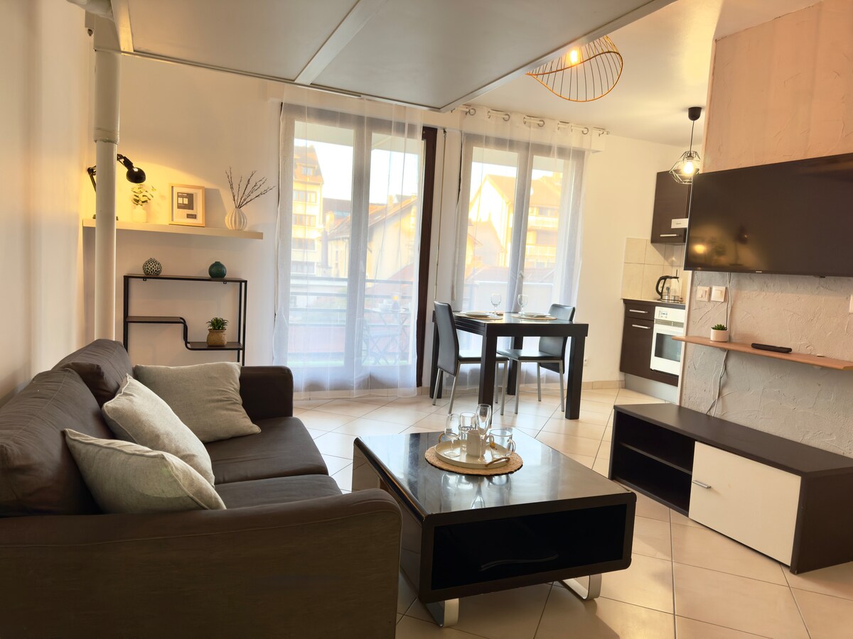 A modern living area features a comfortable dark couch with decorative pillows, complemented by a sleek coffee table. A dining set for two is positioned near a large window, allowing natural light to fill the space. The kitchen is visible, equipped with essential appliances.