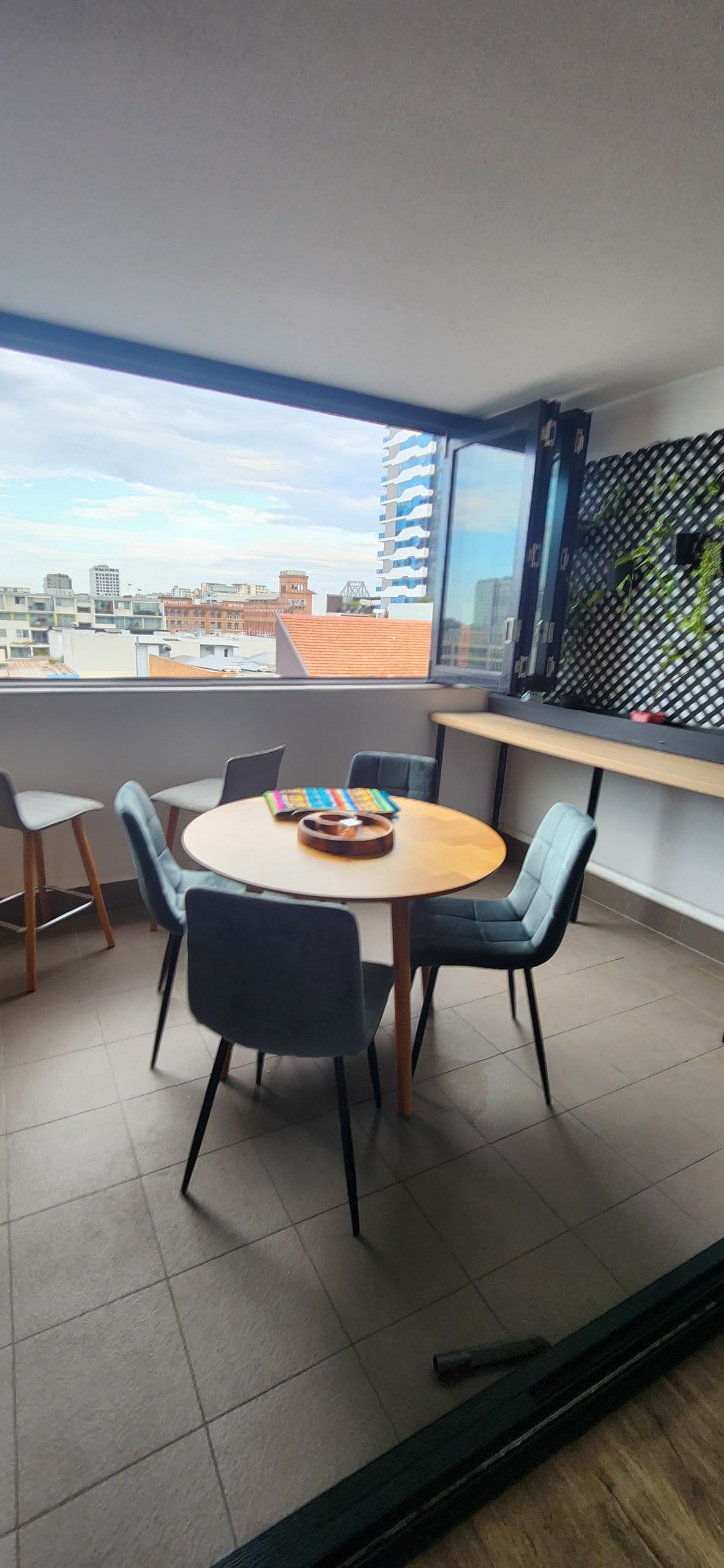 An inviting dining area features a round wooden table surrounded by six upholstered chairs with a view of the city visible through large open windows. A colorful centerpiece decorates the table, enhancing the space's bright and airy feel.