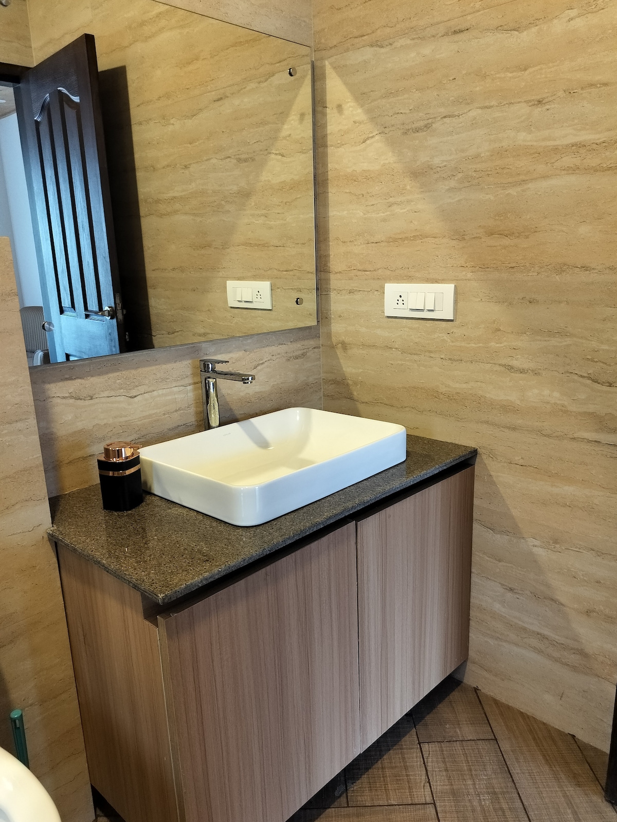 A modern bathroom is equipped with a simple pedestal sink and a rectangular mirror above it. The cabinetry features a light wood finish, complementing the warm tones of the wall tiles. The overall space is well-lit, enhancing its clean and functional design.