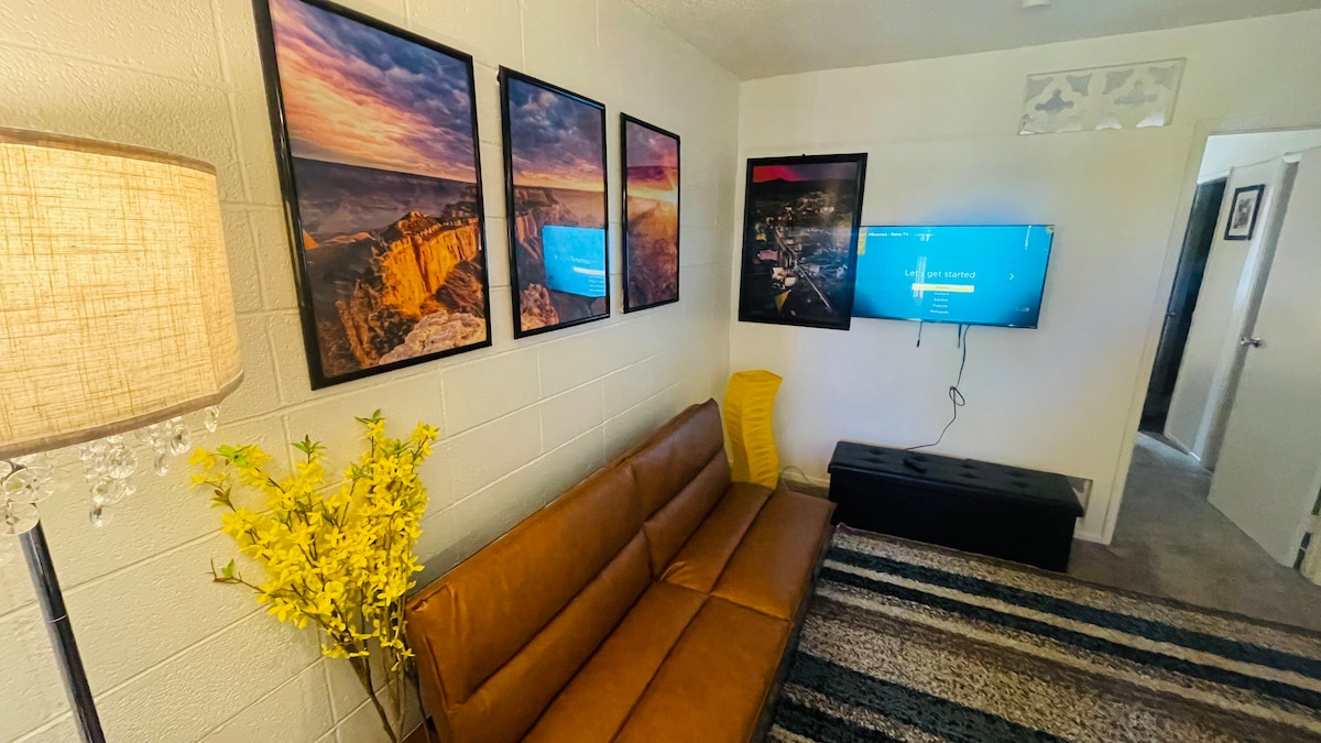 A cozy living area is furnished with a brown couch and a modern coffee table. Vibrant artwork adorns the walls, adding color to the space. A flat-screen TV is mounted nearby, while a lamp and a decorative plant in a vase enhance the ambiance.
