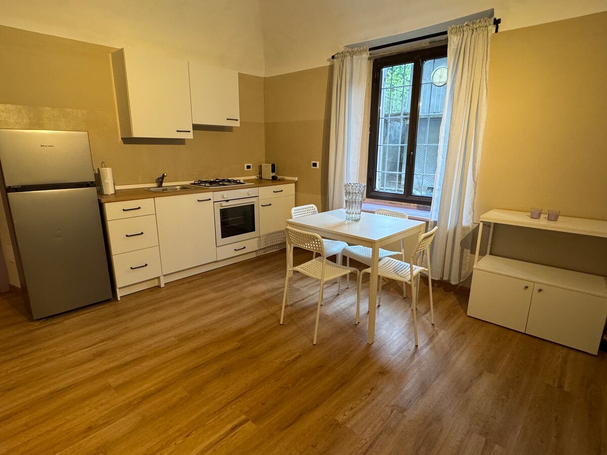 The kitchen area features modern cabinetry and a stainless steel refrigerator. A four-seat dining table made of white material is positioned near a window. Natural light enters through the window, which is framed by light curtains, enhancing the overall openness of the room.